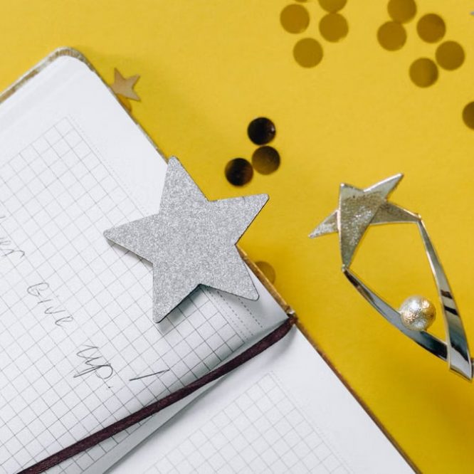 Flat lay of a notebook with motivational text, trophy, and star confetti on a yellow background.